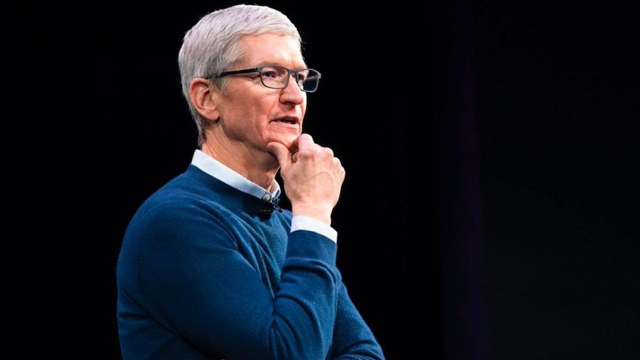 Tim Cook with short gray hair, wearing glasses and a blue sweater, poses thoughtfully against a dark background.